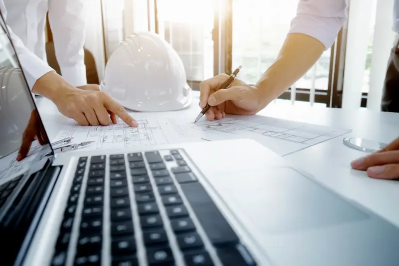 Duas pessoas revisando uma planta ao lado de um notebook e de um capacete